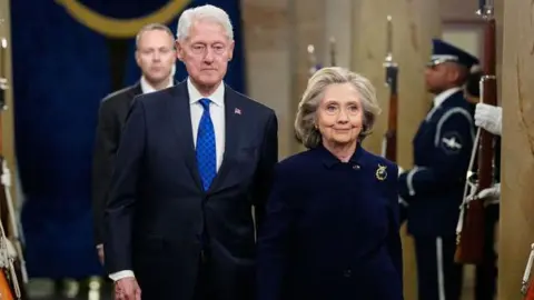 Bill and Hillary Clinton stand walk down a hallway lined with military members holding rifles. The two both wear dark suits