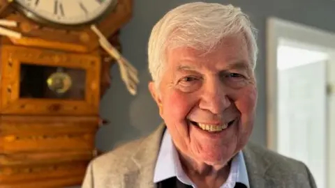 Nigel Thomas is smiling, standing beside a wall-mounted grandfather clock. He has white hair and is wearing a beige suit and pale blue, open-necked shirt. 