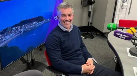 Labour mayor of York and North Yorkshire, David Skaith, is sitting in the BBC Radio York hotseat, waiting to be interviewed by presenter Joanita Musisi. He is smiling at the camera, wearing a blue jumper and an open-neck shirt and has his hands clasped in front of him