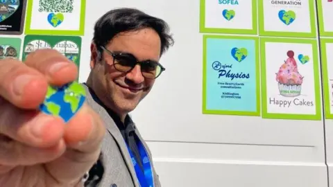 Hearty Earth Hearty Earth ambassador Saman Jamshidifard smiling in front of a white wall with posters displaying different offers from Hearty Earth such as Happy Cakes. He is showing a heart-shaped pin which depicts the earth.