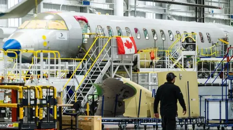 A man walks in front of a Bombardier Global 8000 jet inside a giant warehouse. The jet has a Canadian flag hanging from the windows. 
