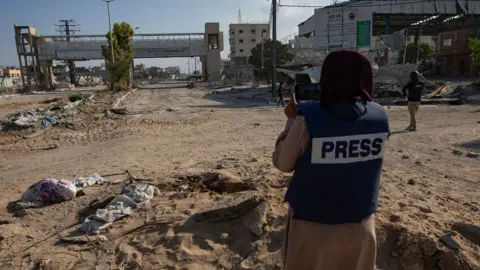 A journalist wearing a protective vest with the word "PRESS" across the back stands on a road strewn with debris in Gaza