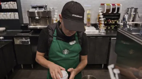 A Starbucks barista offers coffee in New York, wearing a black Starbucks-branded baseball cap and the branded dark green apron, looking down at the coffee cup.