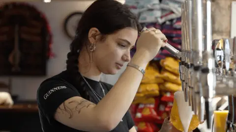 Young white woman with dark hair in a plait, arm tattoos, silver earrings, nose-rings, finger rings and bracelet, pours a pint of larger.  