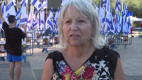 A fair haired lady stands in front of Israeli flags in Hostages Square, Tel Aviv, she is wearing a dark floral top.