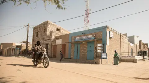 AFP via Getty Images A man rides a motorbike along a dusty road in Timbuktu. Buildings made from concrete blocks line the street