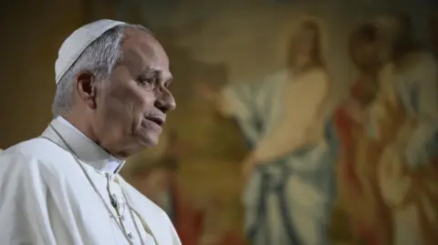 Pope Leo in the Vatican, dressed in papal robes. In front of a big TV showing Americans.