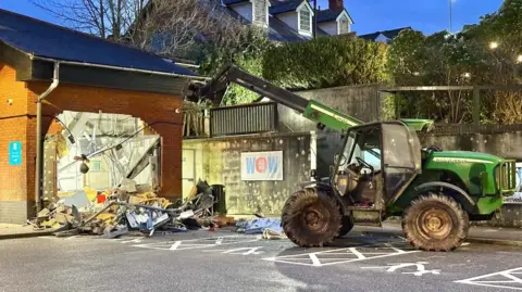 A telehandler is seen next to a damaged brick wall.