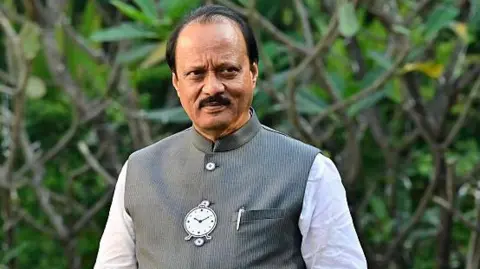 Ajit Pawar poses for a picture at the garden in his residence on January 13, 2026 in Pune. He's wearing a white kurta with a dark grey vest that has the image of a clock and pen in his left pocket