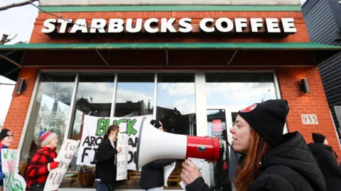 A young female union leader in a black and red woollen hat is standing among other picketers outside a Starbucks. Some of the other picketers in the background are holding signs whose message is not visible in the image. The female union member in the foreground is holding a megaphone as if she is about to start speaking into it.