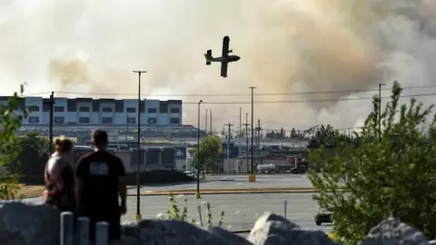 A man and a woman watch a water bomber try to extinguish a wildfire near Bayers Lake at the edge of Halifax, Nova Scotia. Industrial buildings are seen in the background, along with clouds of smoke and the plane. 