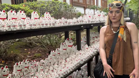 Izabel Rose Izzy, with blonde hair, wearing a brown top and block-patterned bandana stands in front of a shelf containing hundreds of white waving cat figurines