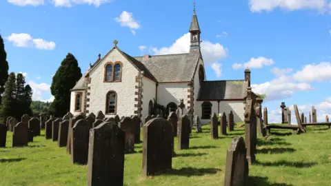 An old church building in white with brick edges and a line of gravestones in front on green grass