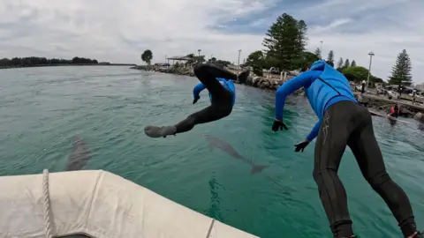 Two people in wetsuits and blue protective equipment dive from the bow of a boat into turquoise water.