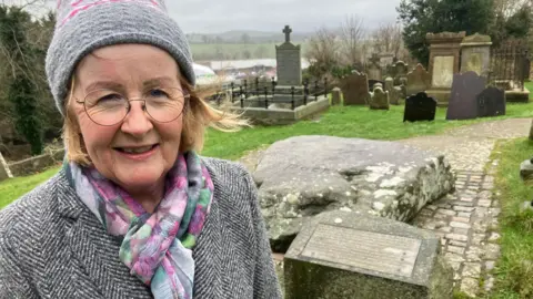 Brigid is wearing a grey coat and colourful scarf, she ha grey hat on her head and a pair of thin glasses. Behind here is a grave yard with headstones, grass and a single cobbled path. 