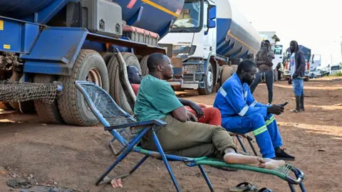 Drivers of Malian tanker trucks sit on chairs beside their vehicles - one dressed in blue overalls checks his mobile phone - as they wait to cross the border between Ivory Coast and Mali - 31 October 2025.