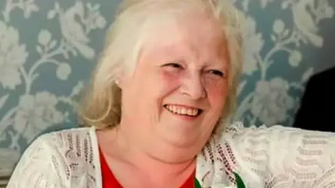 Esther Martin, who is wearing a red top and white cardigan with a flower pinned to it. She is smiling.