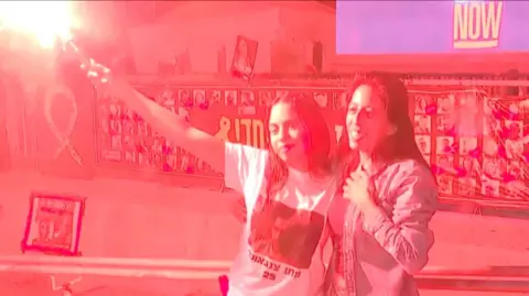 A girl, standing next to her mother, holds a red flare above her head.