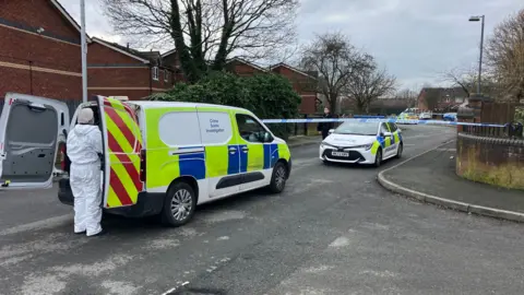 One police car and one police van can be seen on the road, where police tape has blocked off part of the residential street. A person in a full white forensics suit is in the foreground of the picture wearing a mask and head covering. 