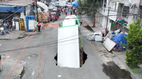 A large white truck is falling into a sinkhole, cordoned off by red tape, on a street in Mexico City.