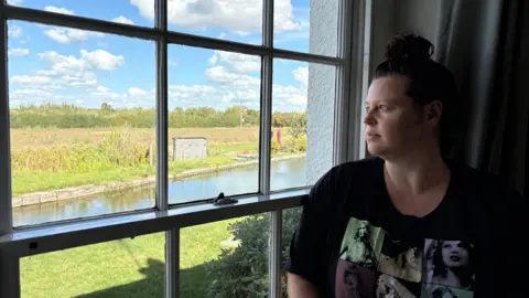 Martin Heath/BBC Kristina Hedley with long brown hair wearing a black T-shirt with green and blue graphics on the front. She is looking through a window made up of several square planes at a canal, with a grass and cereal field with a hedge behind.