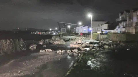 A number of large rocks near the seawall are visible scattered along a footpath.
A number of street lights and buildings can be seen in the background