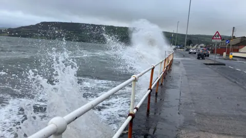 Waves crashing against a sea wall in a village. There are cars travelling down the road.