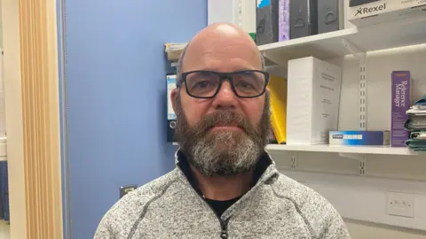 Ellen Knight/BBC Neville pictured looking into the camera. He's wearing a grey quarter-zip fleece and a pair of black-framed glasses. He's pictured indoors inside an office, which has blue walls, and to his left are a set of white shelves which have black, yellow, and white files and folders on them. There's a set of yellow curtains hanging on the left of the image. 