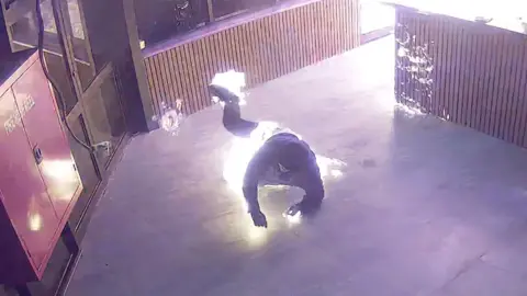 A person wearing black clothes falls on the floor as fire wraps around their body in an empty room.