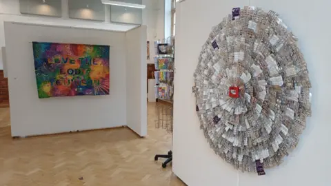 An art exhibition with two large pieces, one is a circle made from layers and layers of dozens of medication blister packs, the other, in the background is a multi-coloured textile work with the words 'Love the body you're in'