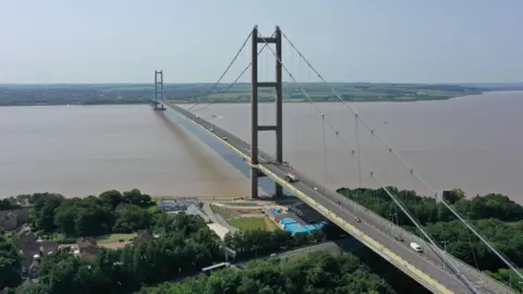 Aerial view of the Humber Bridge showing the bridge spanning the muddy water with vehicles crossing.
