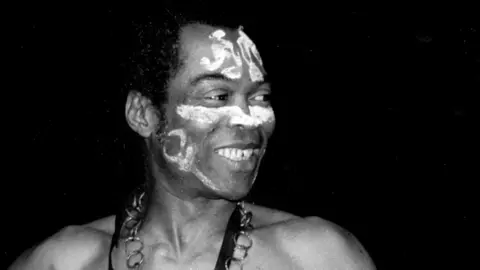 Fela Kuti, bear-chested with white paint on his face and a large chain around his neck, on stage in Detroit, Michigan, in 1986.