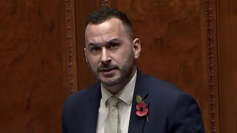 Timothy Gaston speaking in the Stormont Assembly. He has short dark hair, wearing a navy blazer, white shirt and light green tie with a poppy pin on his lapel. Wood panelling is behind him