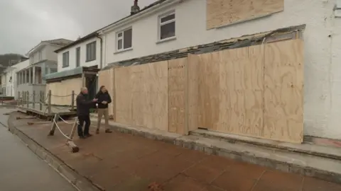 The image shows seafront properties in Torcross, Devon. Several of the homes have windows and doors boarded up after being damaged in Storm Ingrid.