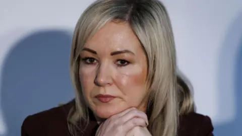 O'Neill has long blonde hair and fringe. She has her hands lapsed below her chin and is staring slightly to her right.