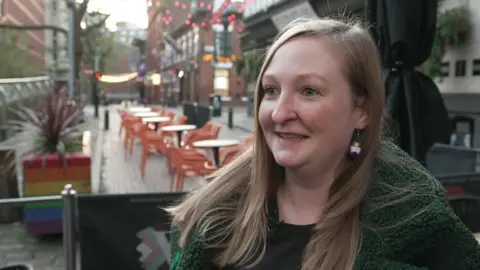 Sarah stands in front of a bar on Manchester's Canal Street, she has long fair hair and wears a green jacket over a black t-shirt
