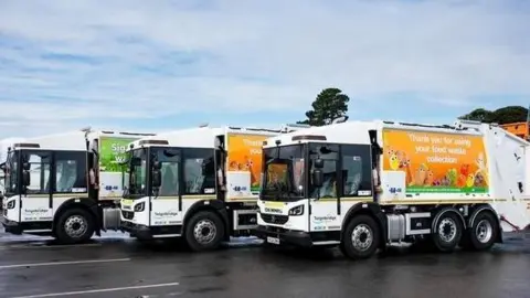 A line of three large refuse vehicles with orange and green banners don the side promoting the use of the local waste collection.