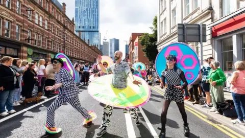 Three participants in a Manchester Pride parade. One wears a black trilby and black and white checked suit (left). The central marcher has a beard, black and white outfit with a multicoloured circle around the midriff while the person on the right has a multi patterned black and white dress  and dyed blue hair.