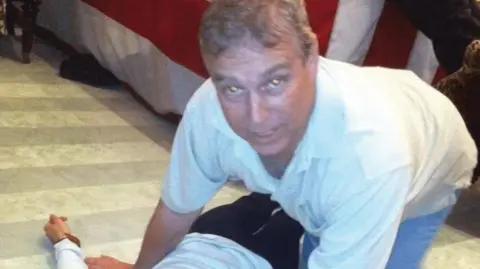 US Department of Justice A grey-haired man dressed in a white polo shirt and blue jeans looks directly at the camera. He is on all fours directly over an unidentified female who is laying on the floor.