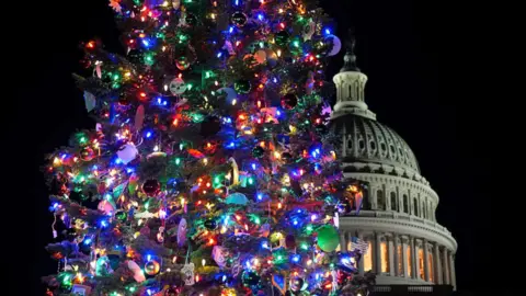 A colorful lit up Christmas tree is in the foreground in a dark background with the US Capitol building behind it.
