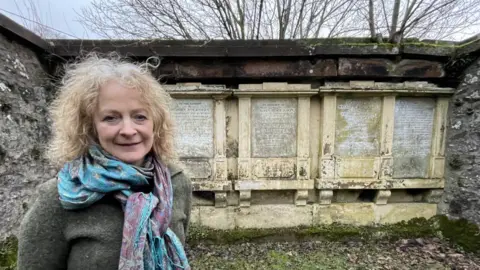 BBC Jan Hogarth, who chairs the Friends of Wamphray Church steering group,  stands in the Rogerson family crypt. She has shoulder length, blonde, wavy hair and is wearing a dark green jumper and a large blue and purple silk scarf. 