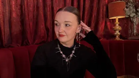 A woman wearing a black top, a necklace, and earings sits in a red booth, with red curtains in the background. She is looking to the left of the camera