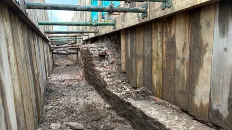 Part of a medieval brick wall can be seen in a trench dug beneath a road. It is made of red bricks - though they look brown due to the soil. The surviving wall is quite low, though it builds up to a higher part, with about 14 layers of brick. The trench is protected by temporary wooden structures on either side, with green metal poles braced between them.