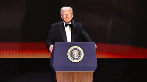 Trump in a suit and tie, stands on stage before a POTUS lectern.