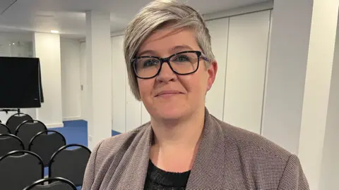 Deputy Haley Camp - A woman with mid-length grey hair, wearing a checked jacket and a black blouse. 