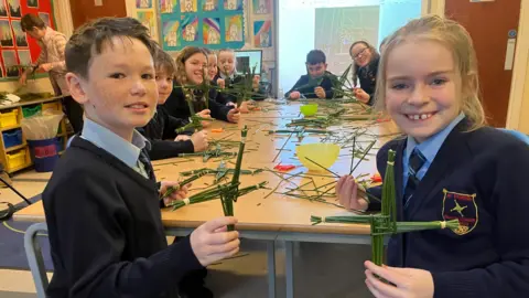 Two school pupils are holding a green cross. They are wearing blue shirts with navy ties and jumpers. There are more children holding crosses in the background. They are inside a classroom. 