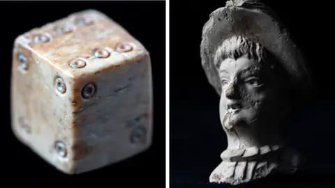 A close up a medieval dice and part of an imported French pipe with the moulding of a woman's head found during the HS2 archaeological excavations