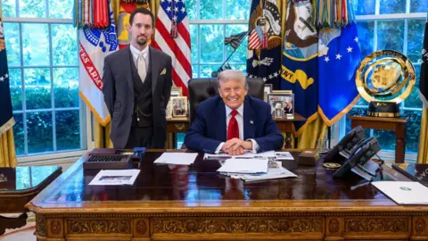 Trump sits behind desk in blue suit and red tie while Antoni stands in three-piece gray suit with a beige tie, white collar and watch chain in front of windows covered in colorful flags and banners