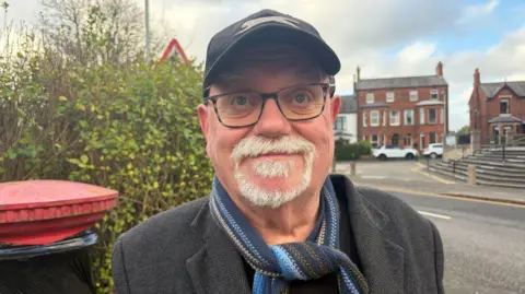Mid-shot of a man with a cap, glasses and a gray beard. He has a blue scarf and a grey jacket. Behind him, to his left is a postbox, wrapped in black except for its top. He is standing next to the road