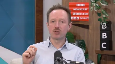 A man, Adam Fleming in the Newscast studio. He has short hair and wears a white shirt